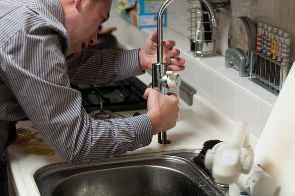 A plumber crouching under a kitchen sink with tools, writing an estimate on a clipboard — professional, realistic, and relatable for homeowners researching costs.